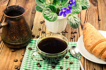 Cup of black coffee on green napkin with croissant, turkish coffee pot, flower pot, wooden table in cafe