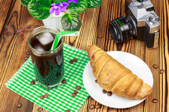 Glassful of black coffee with ice and tubule on green napkin. croissant, vintage camera, flowerpot, wooden table