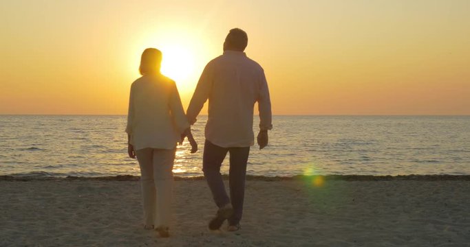 Steadicam shot of senior couple coming to the beach to see the beauty of sunset. They embracing and looking at sea and sundown