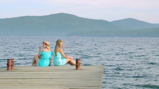 Cute girls with wreaths on their heads are resting on the river. Two young beautiful woman in a blue dress sits on a wooden pier on the river.