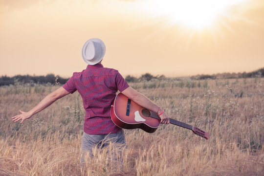 Young retro styled man with acoustic guitar in wheat field looking at sun to find inspiration for the next song. Music, art and lifestyle concepts.  