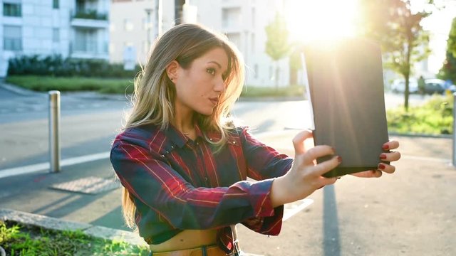Young beautiful caucasian blonde hair woman outdoor in city back light hlding a tablet taking selfie &ndash; vanity, technology, social network concept
