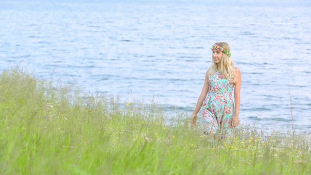 Beautiful girl walking in meadow wearing floral wreath. Young adult. Wearing stylish dress with flower print.