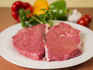 Two pieces of raw beef on a plate and some vegetables in background.