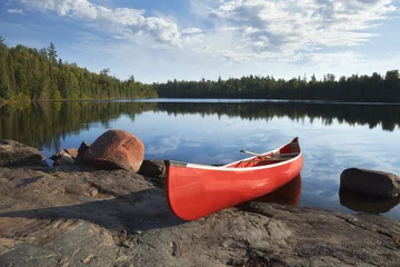 Foto op Canvas Red canoe on rocky shore of calm lake with pine trees © Daniel Thornberg