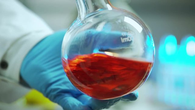 The gloved hand of a laboratory chemist swirls a flask containing a red chemical.