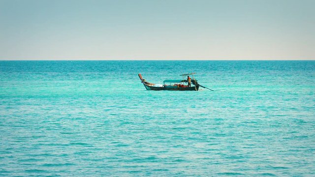 Extreme zoom shot of a longtail boat, cruising along the bold, blue horizon, over a tropical sea.
