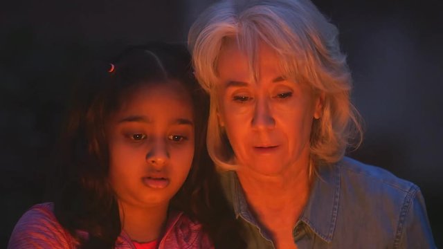 A grandmother shows her young granddaughter how to roast a marshmallow over an open fire.