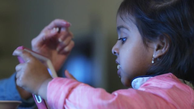 This small girl continues to watch her IPAD as she gets a bite of food fed to her like a little bird.