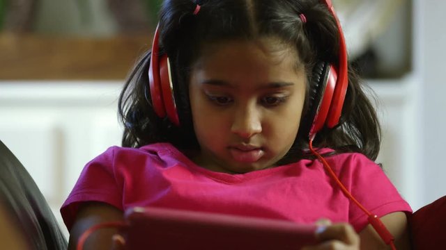 A pretty Indian child with headphones on looks up briefly and smiles before returning to entertainment on her tablet pc.