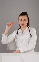 Beautiful nurse in white robe and stethoscope is making her report at the white table against of grey background
