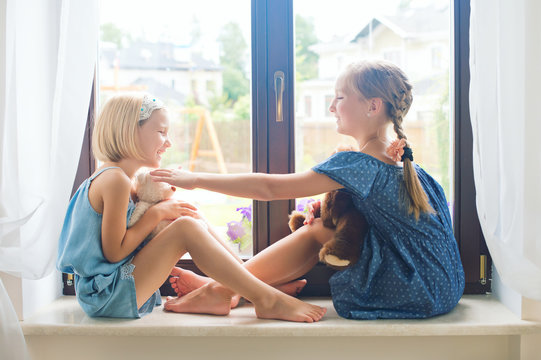 Two cute european toddler girls sitting on sill near window at home playing toys