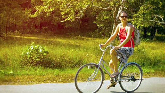 FullHD video - Adventurous tourist riding a rented bicycle along a rural road with a green, grassy field and beautiful, wild trees in the background.