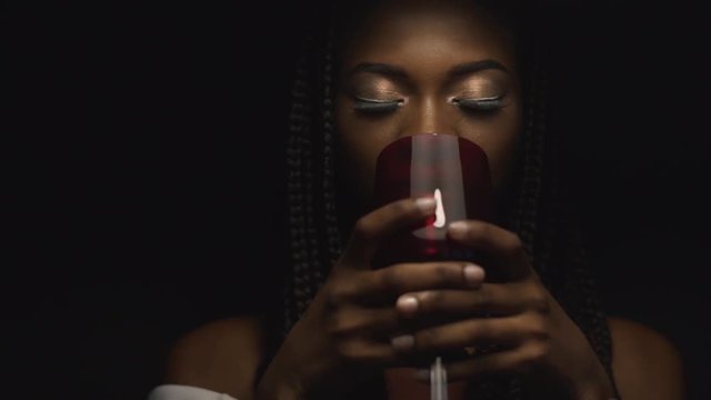 Big close-up of sexy mysterious african american female model drinking a glass of red wine on black background in slow-motion