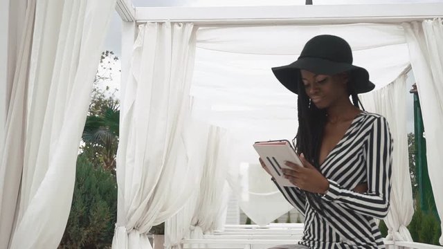 Glamorous african american lady in a striped cocktail dress and black hat using her tablet computer on the luxury hotel outdoor background