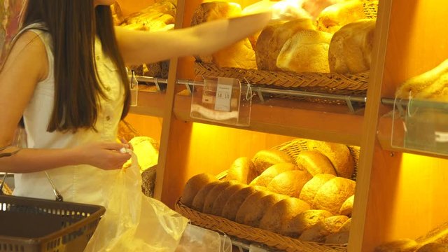 Woman chooses and putting a fresh bread loaf into a package in the supermarket. Young girl taking a loaf of bread from the shelf and putting it into the basket. Shopping in the grocery