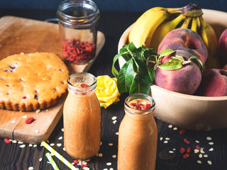 Pumpkin, banana and peach vegan smoothie with soy milk, sesame seeds, oatmeal flakes and goji berries on dark wooden table with vegan spelt cake and bowl with fruit. Healthy breakfast still life
