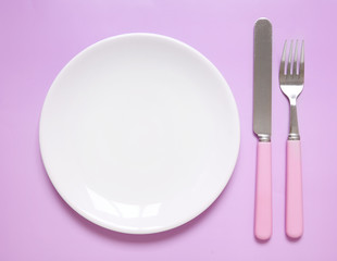 An empty dinner plate, knife and fork on a pink background, arranged as a place setting