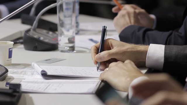 Man's hand writes notes in a very important point. International Forum, a businessman report on the economic theme. People sit at the table.