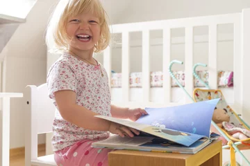 Fotobehang Kleinkind im Kinderzimmer schaut lachend ein Buch an. © mmphoto