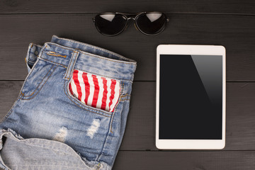 Summer women's accessories: shorts, tablet, sunglasses on wood background.