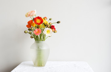 Red, white, yellow and orange poppies in a glass vase on table with a white tablecloth