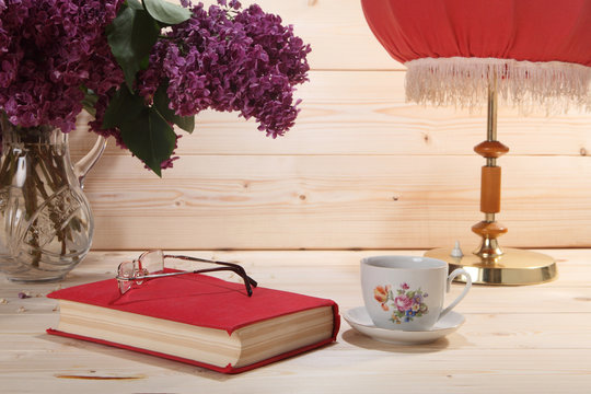 Bouquet of lilacs, book, spectacles, cup of tea and table lamp o