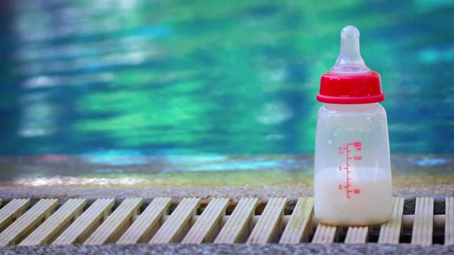 Four ounce baby bottle, half full of milk, with a silicone nipple, sitting on the overflow drainage at the edge of a swimming pool.
