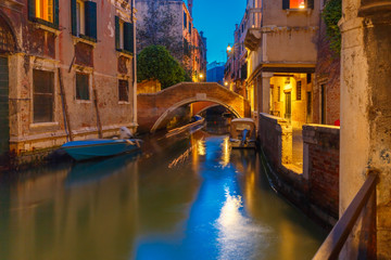 Lateral canal and pedestrian bridge in Venice at night with street light illuminating bridge and houses, with docked boats, Italy