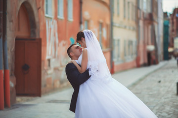 elegant groom lifting up his beautiful brunette bride on the background of old white wall