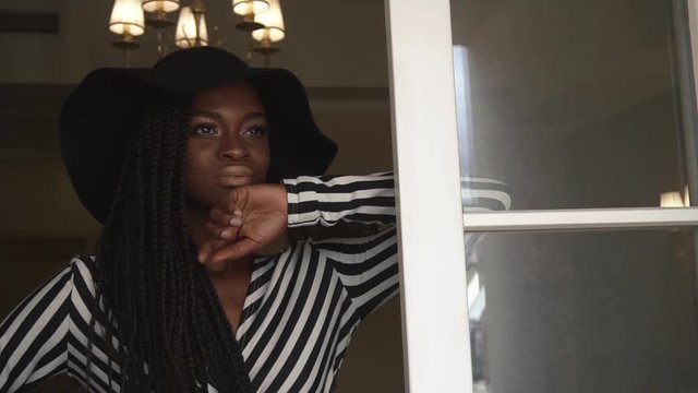 Close-up pretty face of black african lady in black hat and striped cocktail dress posing at her luxury apartments background