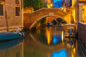 Fototapeta na wymiar Lateral canal and pedestrian bridge in Venice at night with street light illuminating bridge and houses, with docked boats, Italy