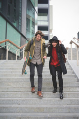 Obraz premium Young couple of caucasian woman and man, with skate and moustache, walking down the stairs hugging and smiling - youth, carefree, freshness concept