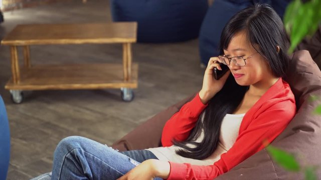 Girl with long black hair sitting in cafe talk on mobile phone. On her young beautiful face embarrassed smile. Vietnamese model wearing casual ripped jeans and stylish cardigan. She has poor eyesight