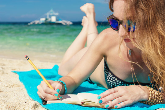 young european woman with sunglasses is lying on the coast of tropical turquoise sea and wrigting by pencil in notepad at sunny summer day