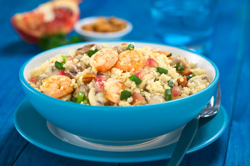 Couscousgericht mit gebratenen Garnelen, Champignons, Mandeln, Granatapfel und Frühlingszwiebeln in blauer Schüssel (Selektiver Fokus, Fokus auf die Vorderseite der oberen Garnelen)