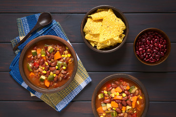 Veganischer Chili aus Kidneybohne, Karotte, Zucchini, Paprika, Mais, Tomate, Zwiebel und Knoblauch, mit Maischips und rohe Bohnen an der Seite, fotografiert mit natürlichem Licht