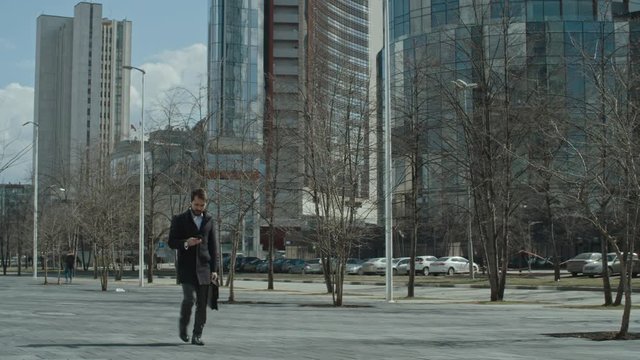 Businessman using his smart phone and answering a call while walking on the city street; modern glass office buildings in the background