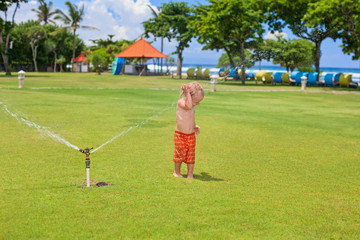 Fototapeta premium Happy baby boy play with sprinkler spray, swim and splash with fun under water drops on green grass lawn. Travel, active lifestyle, parenting, people activity on summer beach holiday with children.