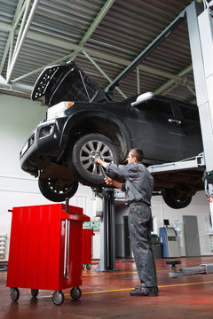 Male Mechanic Fixing Wheel On Raised SUV Car In Garage. Young Man In Mechanic Uniform Works With Wheel Of Raised On Lift Car In Auto Service Station. Mechanic Doing Service Maintenance For SUV