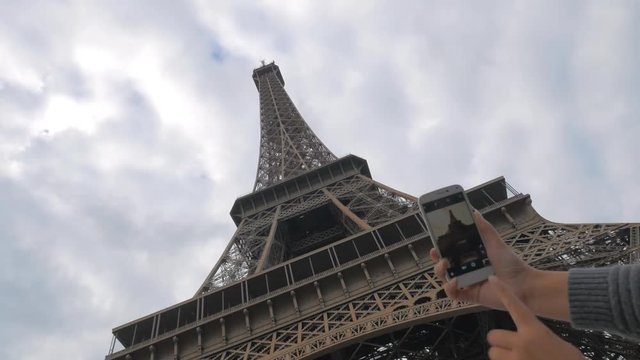 Low angle shot of woman tourist using smart phone to make a photo of Eiffel Tower against cloudy sky