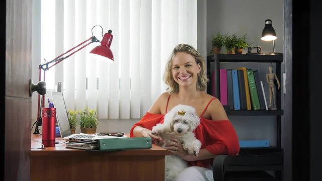 People, pets and love for animals. Woman at home in office room works on laptop holding her little dog. She turns to the camera smiling. Slow motion