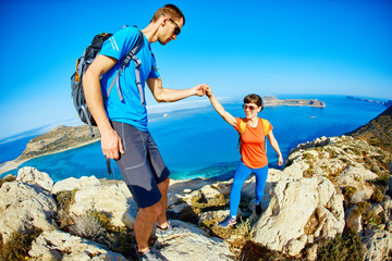 couple of travelers with backpack standing on the cliff against sea and blue sky at early morning. Balos beach on background, Crete, Greece. a man helping a woman climb on the rock extends her hand