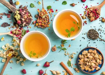 Foto auf Acrylglas Tee Zwei Tassen gesunder Kräutertee mit Minze, Zimt, getrockneten Rosen- und Kamillenblüten in Löffeln auf blauem Hintergrund