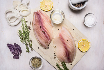 raw fillet of tilapia with spices and herbs, lemon and pepper on a cutting board on a white background rustic village top view