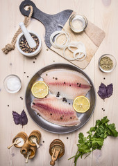 raw fillet of tilapia with spices and herbs, lemon and pepper, onion, parsley, rosemary on a pan on a white background rustic background top view