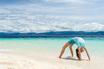 Fototapeta na wymiar young slim teen girl do gymnastic exercise at white sand beach of tropical sea under blue sky