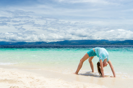 young slim teen girl do gymnastic exercise at white sand beach of tropical sea under blue sky