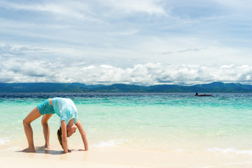 Fototapeta na wymiar young slim teen girl do gymnastic exercise at white sand beach of tropical sea under blue sky