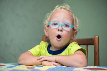 Удивленный ребенок в очках с открытым ртом сидит за столом 